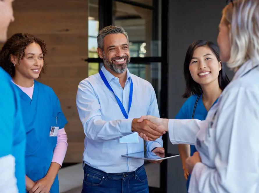 A group of healthcare professionals stands together indoors. A man in business casual attire from a healthcare marketing agency shakes hands with a woman in a white lab coat while two nurses in blue scrubs smile nearby. The atmosphere appears friendly and collaborative.