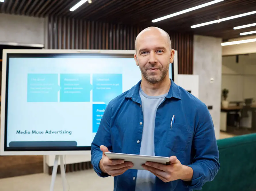 A bald man in a blue shirt holds a tablet and smiles at the camera. He stands in a modern office with wood and glass elements, where a large screen behind him displays a healthcare digital marketing agency presentation slide.