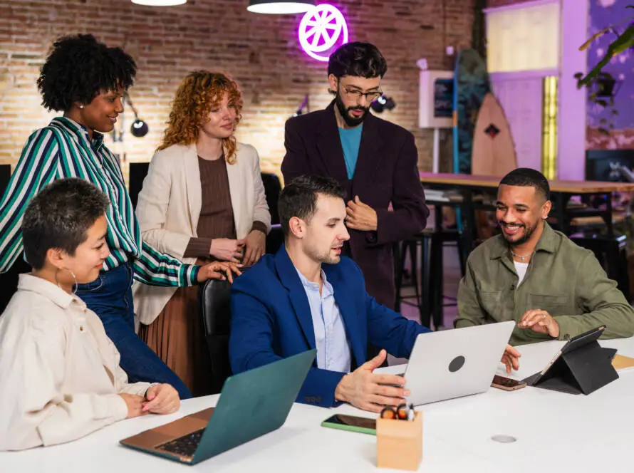 Six people gather around a table with laptops and tablets in a modern office space. One man in a blue suit gestures while talking; others listen and smile, suggesting teamwork at a healthcare digital marketing agency. Exposed brick walls and vibrant lighting complete the room.