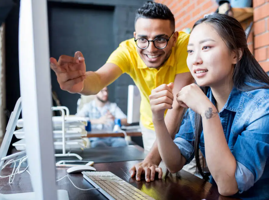 A smiling man in a yellow shirt points at a computer screen, assisting a focused woman in a denim shirt. They collaborate on digital marketing healthcare strategies in a modern office with exposed brick and shelves in the background.