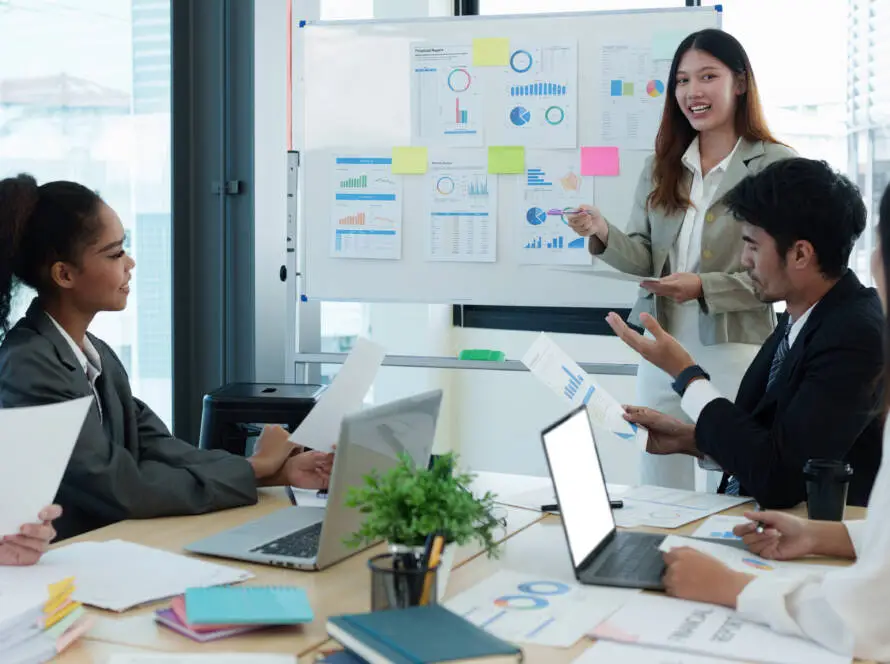 A woman stands by a whiteboard with charts and sticky notes, presenting to four colleagues at a healthcare marketing agency. Laptops, documents, and a small plant sit on the table in their modern office with large windows as everyone engages in discussion.