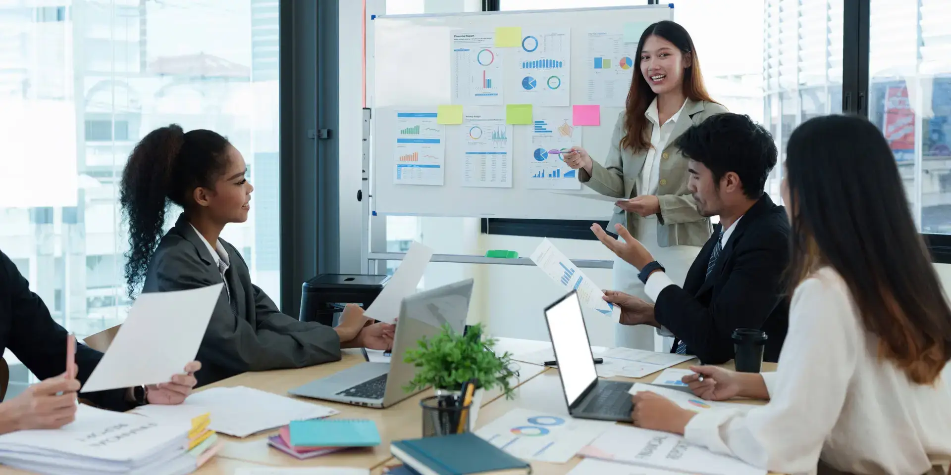 A woman stands by a whiteboard with charts and sticky notes, presenting to four colleagues at a healthcare marketing agency. Laptops, documents, and a small plant sit on the table in their modern office with large windows as everyone engages in discussion.