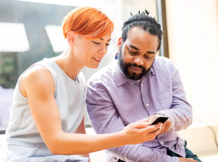 A woman with short red hair and a man in a lavender shirt sit close together indoors, both looking at a mobile phone. They appear engaged, possibly exploring options from a healthcare digital marketing agency on the phone.