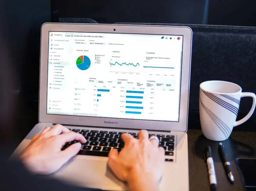 A person types on a MacBook laptop displaying data analytics with graphs, charts, and tables for healthcare SEO. A coffee mug and pen rest nearby. The screen shows blue, green, and red charts representing business statistics.