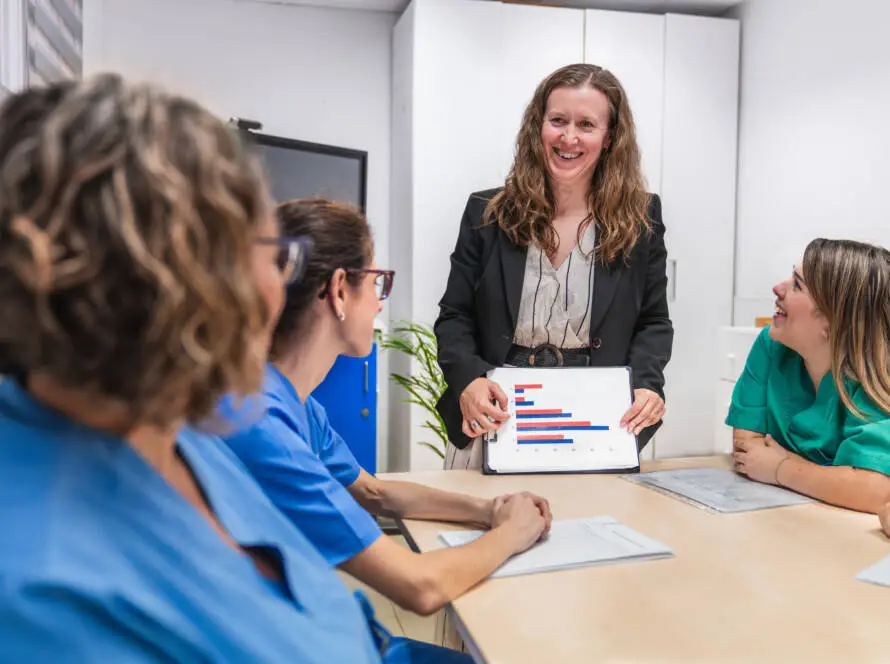 A healthcare SEO consultant in a suit stands at a table, smiling and showing a bar chart on a tablet to three women in scrubs who are listening and taking notes in a brightly lit meeting room.