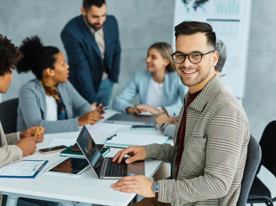 A smiling man with glasses, a healthcare SEO consultant, sits at a table using a laptop in a modern office. Four colleagues talk and share documents in the bright setting, with charts on a board and digital marketing healthcare materials on the table.