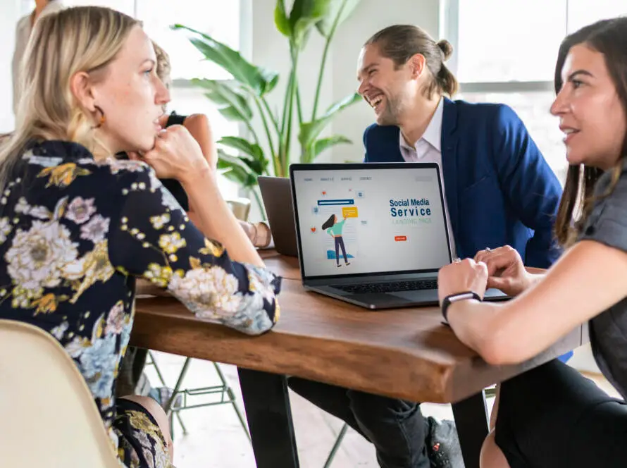 Three people sit around a wooden table, talking and smiling. A laptop displays a “Social Media Service” website, ideal for a medical marketing agency focused on healthcare SEO. Large green plants and big windows suggest a bright, modern office.