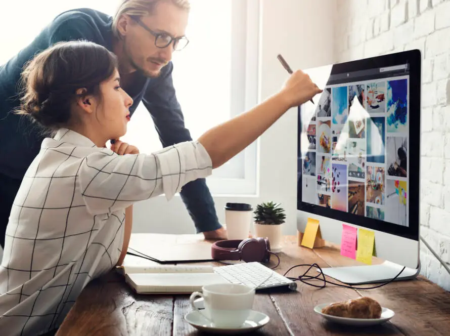 A woman sits at a wooden desk, pointing at a computer screen filled with colourful images, while a man beside her observes. The workspace, dotted with coffee and notes, hints at digital marketing healthcare or healthcare SEO strategies in action.
