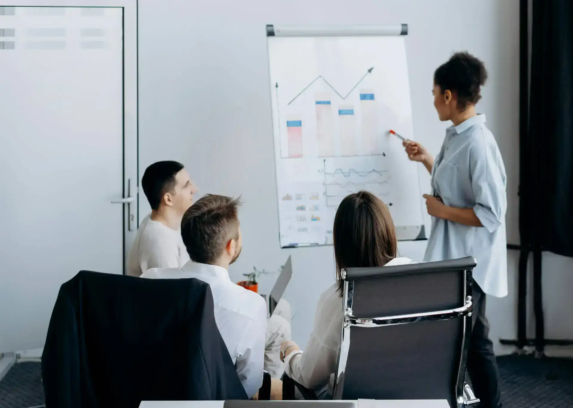A woman stands by a flip chart, pointing at graphs with a red marker as three colleagues from a healthcare digital marketing agency listen closely. The modern office setting features charts with bars and line graphs.
