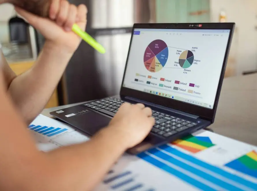 A person holds a highlighter and types on a laptop displaying colourful pie charts and bar graphs. Various printed charts are spread on the table, highlighting in-depth analysis typical for a medical marketing agency or digital marketing healthcare firm.
