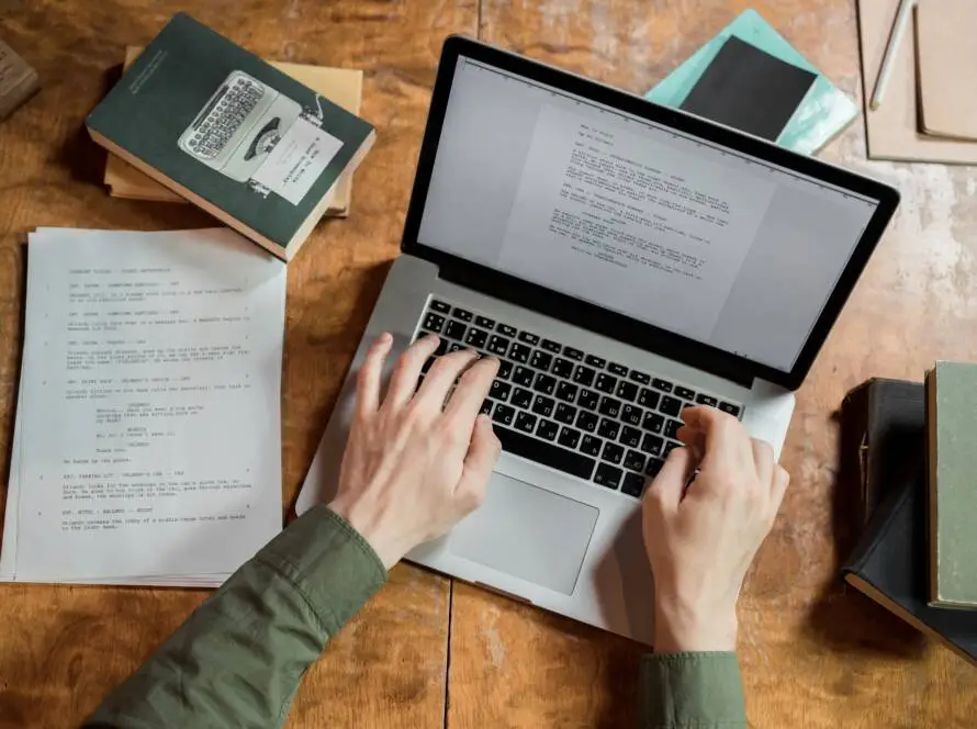 A person types on a laptop at a wooden desk cluttered with books, papers, and a notebook. The screen displays text, hinting at writing or editing work for a healthcare SEO consultant. The person wears a green long-sleeved shirt.