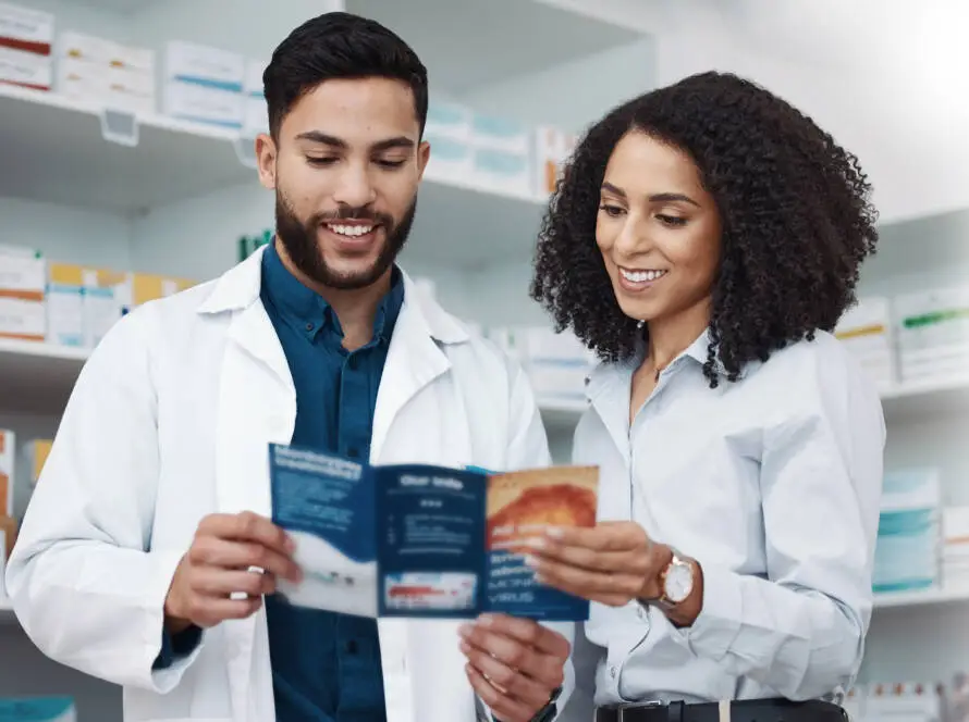 Two people stand in a chemist, smiling whilst reading a blue informational leaflet together. One wears a white lab coat and the other a light blue shirt. Shelves with various medicines are visible, highlighting effective strategies from a medical marketing agency.