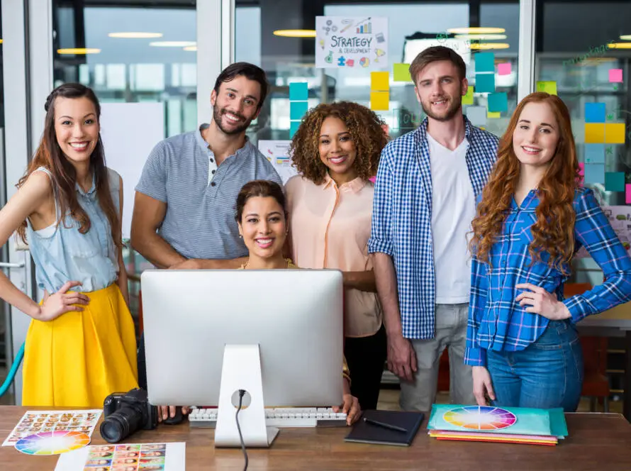 Six young adults, three women standing, one man standing, and two women seated by a large monitor, smile at the camera in a modern medical marketing agency office with glass walls, colourful Post-it notes, charts, and a camera on the desk.