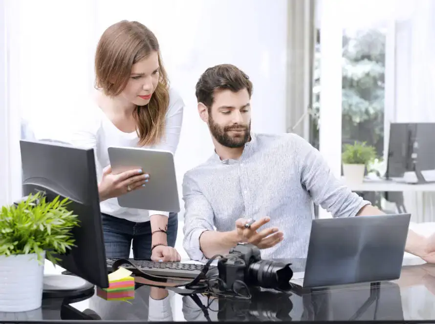 A woman holding a tablet stands next to a seated man using a laptop as they discuss work for their healthcare digital marketing agency. A camera, plant, keyboard, and sticky notes are on the desk in a bright, modern office setting.
