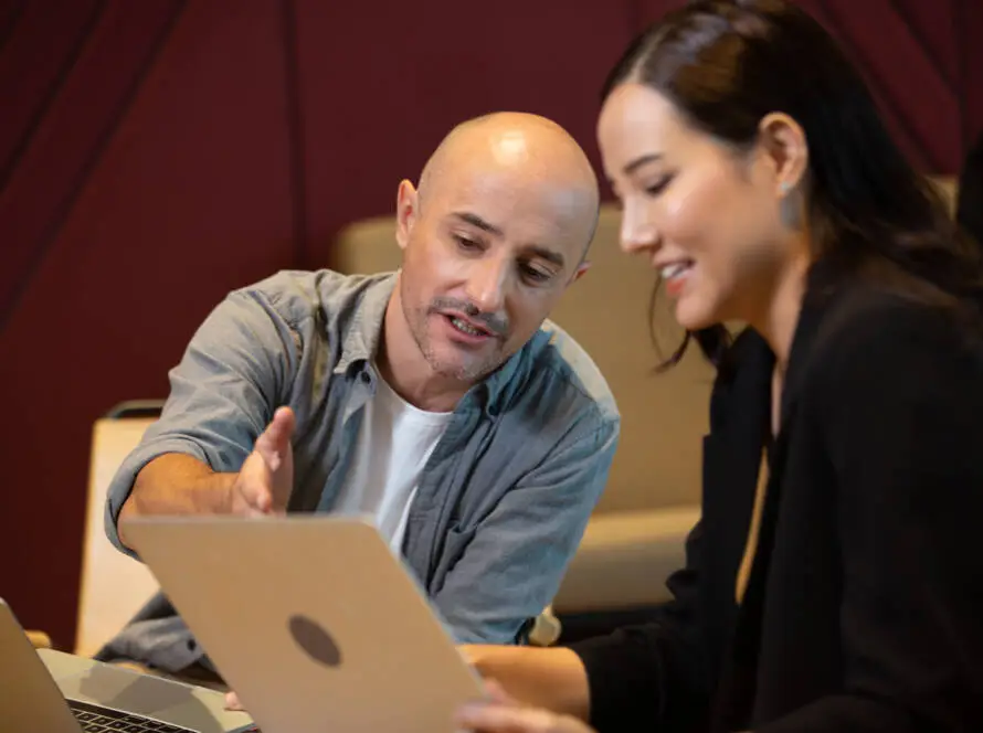 A man and a woman sit together at a table, both looking at a laptop screen. The man gestures whilst speaking about digital marketing healthcare, and the woman smiles, suggesting an engaged and collaborative work environment.