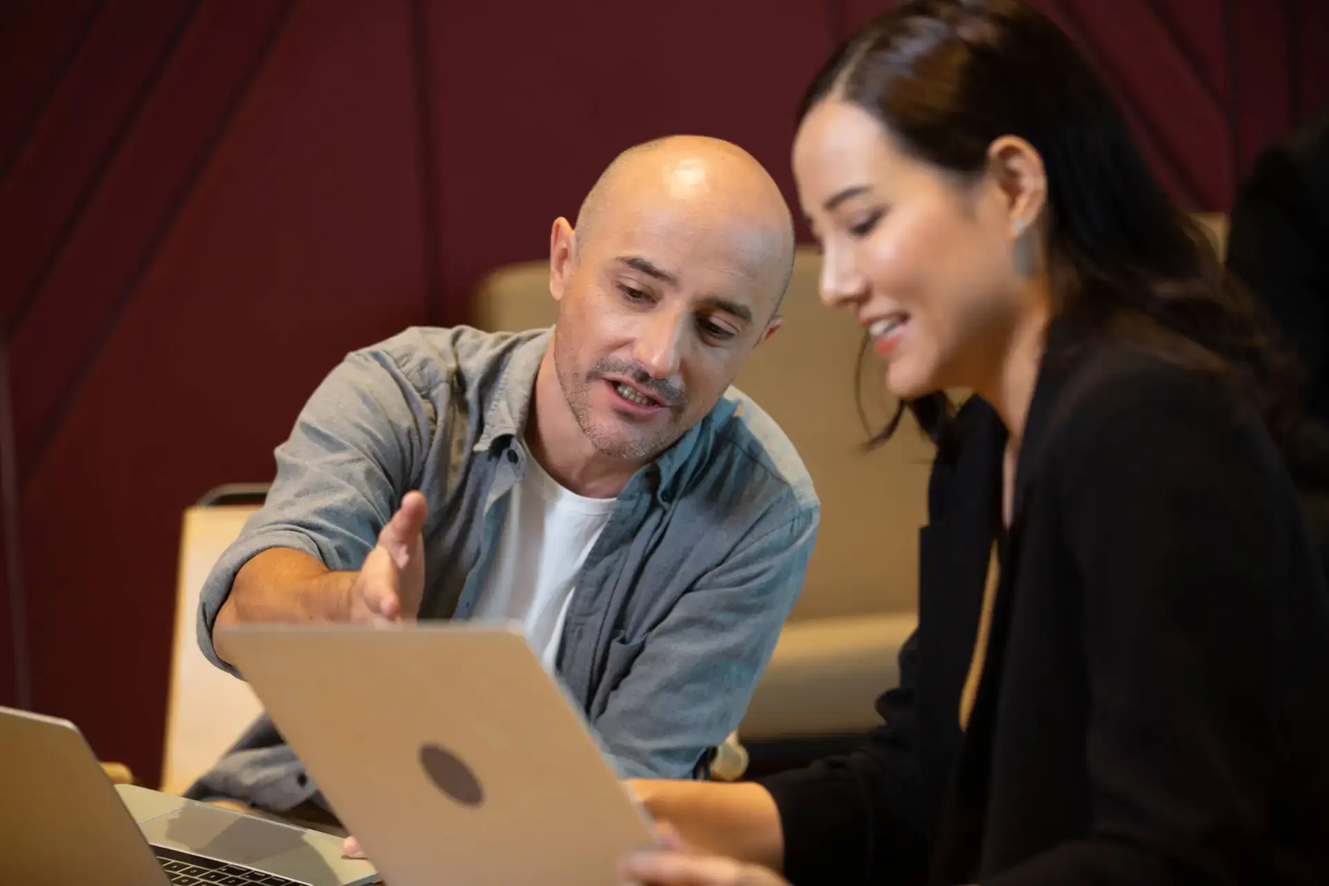 A man and a woman sit together at a table, both looking at a laptop screen. The man gestures whilst speaking about digital marketing healthcare, and the woman smiles, suggesting an engaged and collaborative work environment.