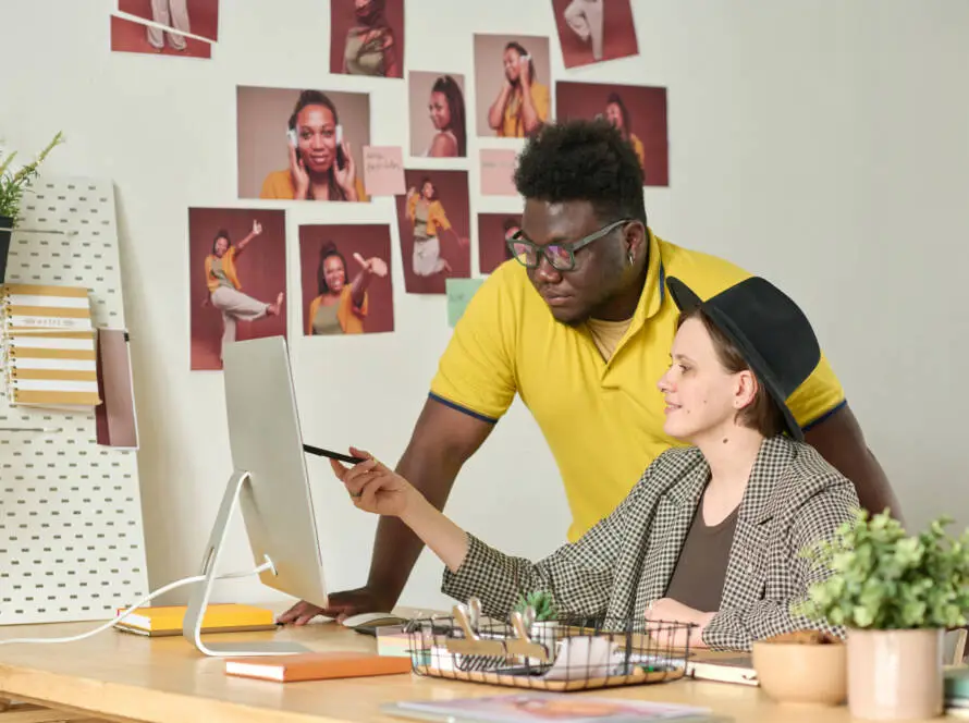 Two colleagues at a medical marketing agency collaborate at a computer. One stands in a yellow shirt and glasses, while the other, in a checked jacket and hat, sits and points at the screen. Portrait photos decorate the wall behind them.