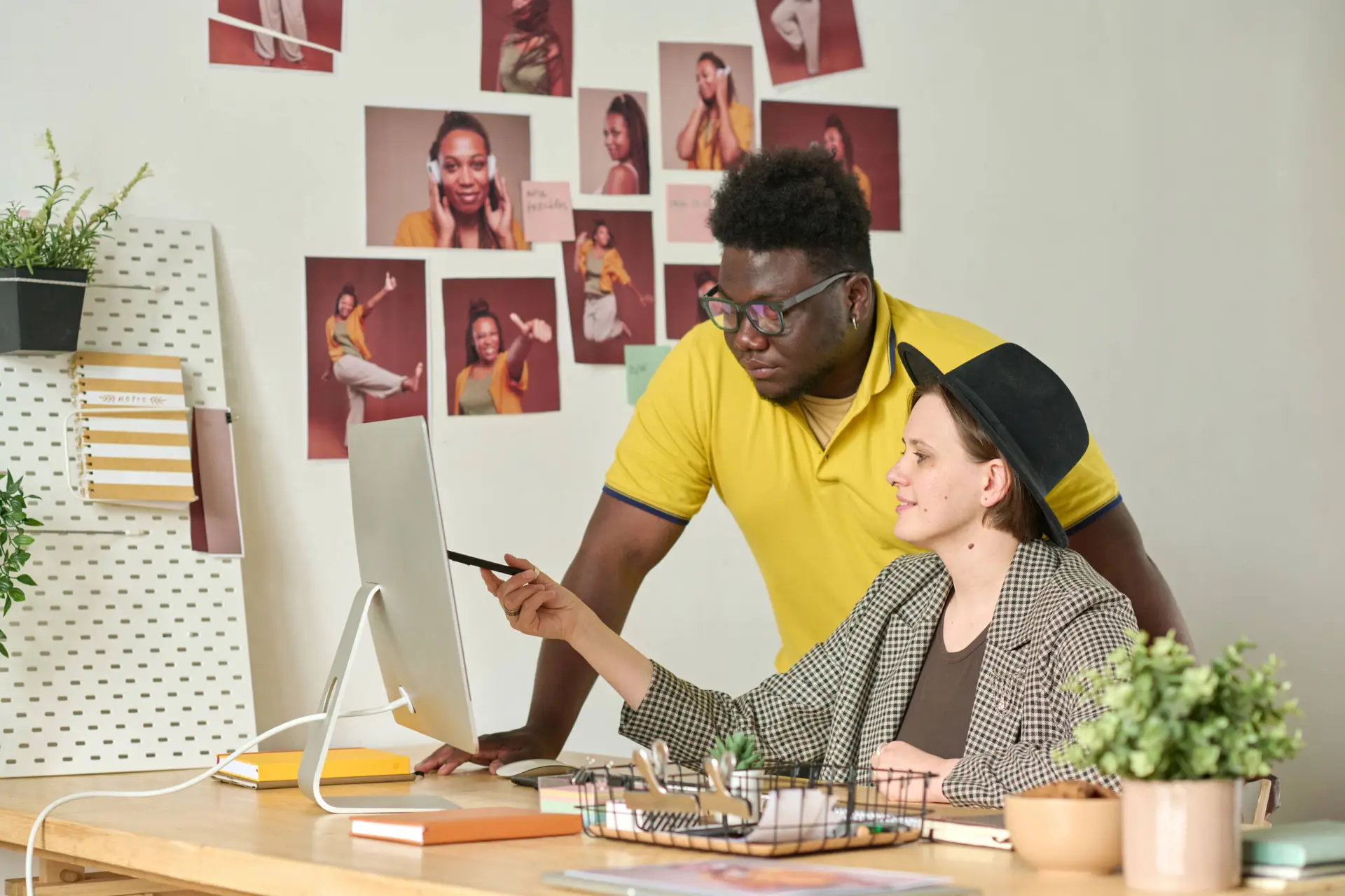 Two colleagues at a healthcare digital marketing agency collaborate at a desk. One points at a computer screen with a pen whilst the other leans in. The wall is decorated with photos, and office supplies and plants fill the desk.