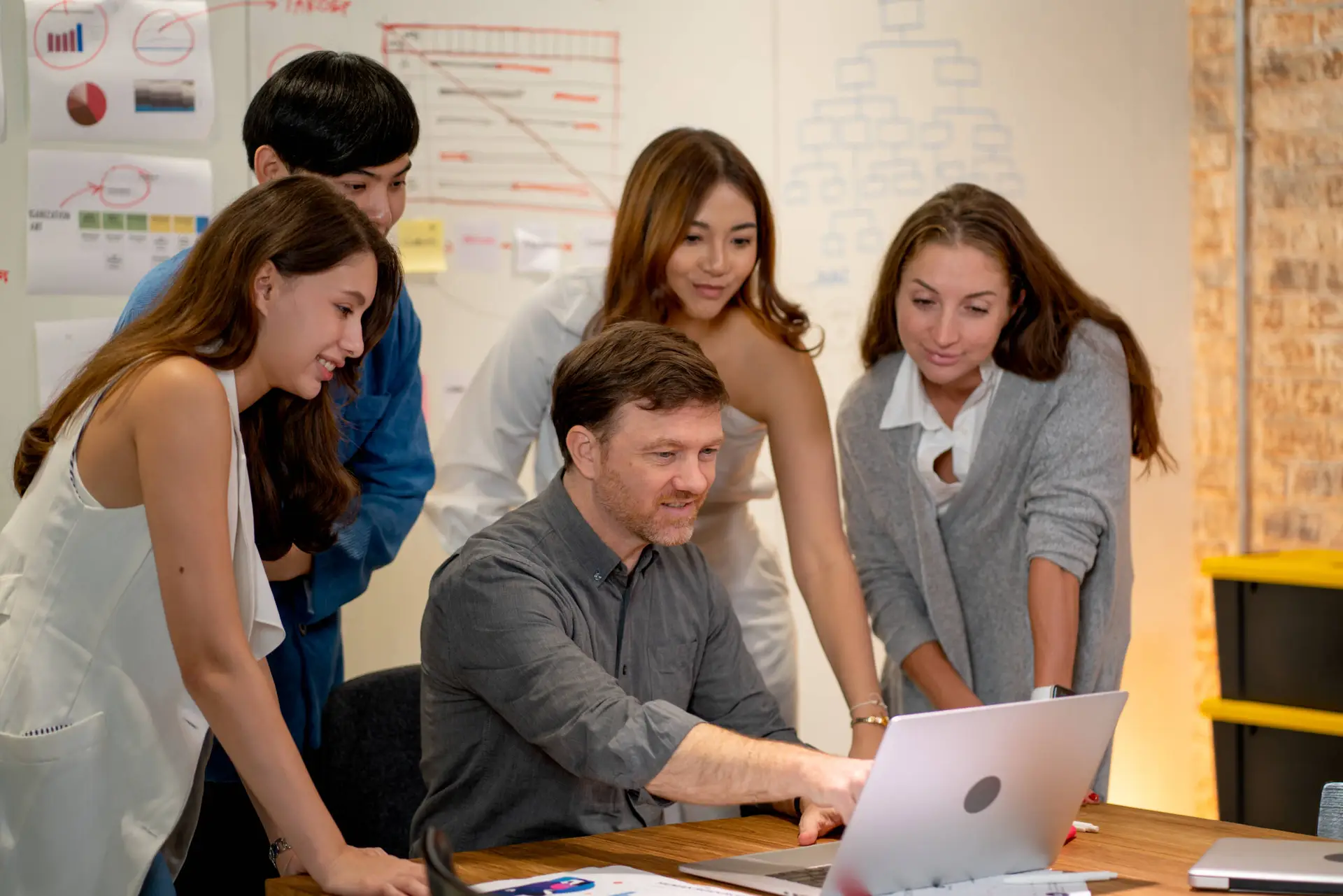 Five colleagues, four women and one man, gather around a desk in a modern office. The man points at a laptop screen while others stand closely, smiling and engaged—brainstorming strategies for their healthcare digital marketing agency. Charts line the wall behind them.