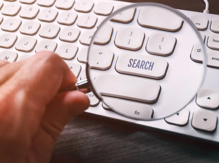 A hand holds a magnifying glass over a white computer keyboard, enlarging the SEARCH key—symbolising how a healthcare digital marketing agency helps clients find and boost online visibility in the medical field.