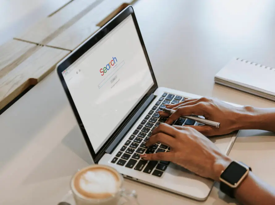 A person types on a laptop with a search engine open, researching healthcare SEO. Nearby are a mobile phone, notebook, pen, and coffee. The person wears a smartwatch and sits at a clean, light-coloured workspace.