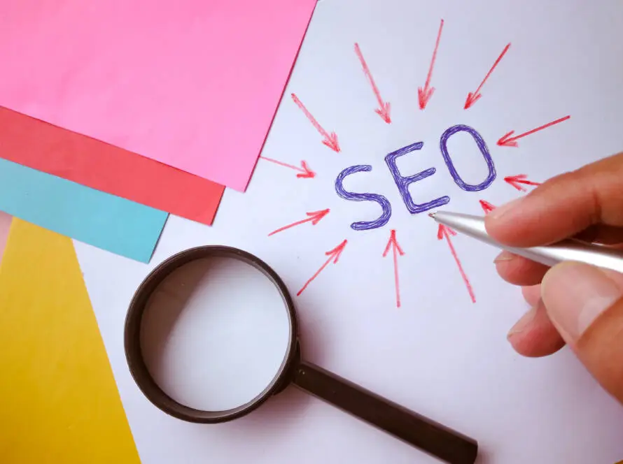 A hand holding a silver pen points to the word “SEO” written in blue on white paper, surrounded by red arrows. A black magnifying glass rests nearby, highlighting the focus of a healthcare SEO or healthcare digital marketing agency. Colourful papers are in the background.