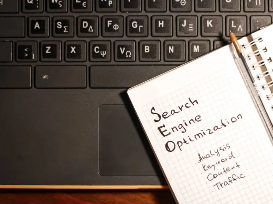 A black laptop keyboard with Greek letters sits next to an open notebook. The page reads “Search Engine Optimisation (SEO)” with handwritten notes: “Analysis, Keyword, Content, Traffic.” Perfect for a healthcare SEO consultant brainstorming strategies.