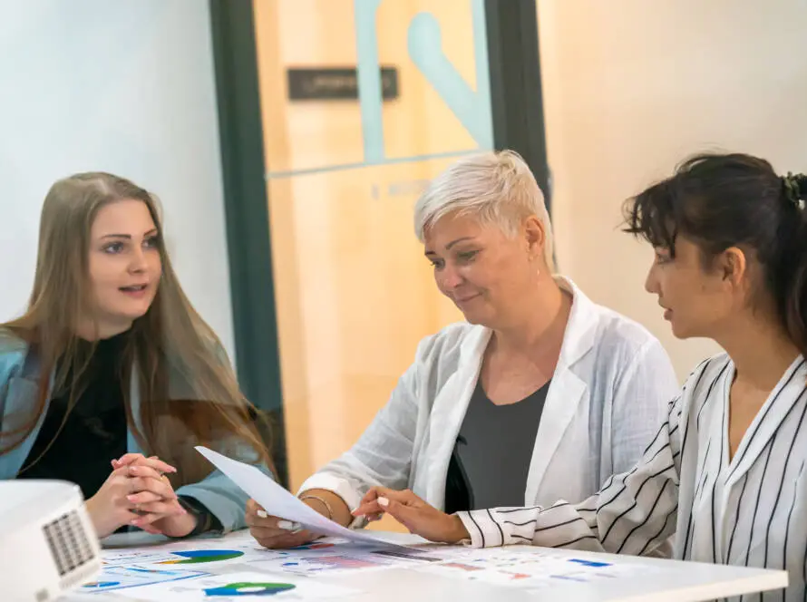 Three women sit at a table in a modern office, engaged in a collaborative business meeting for a healthcare digital marketing agency, discussing documents and charts and sharing insights to drive success.
