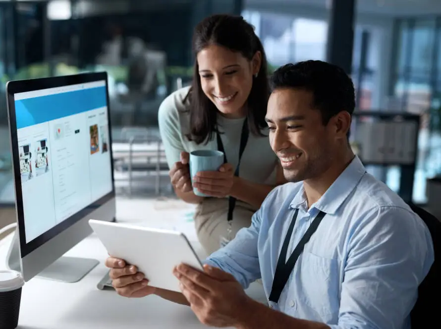 A man sits at a desk holding a tablet and smiling, while a woman with a blue mug stands beside him. They appear to be collaborating in a modern office, possibly working with a healthcare digital marketing agency on an exciting project.