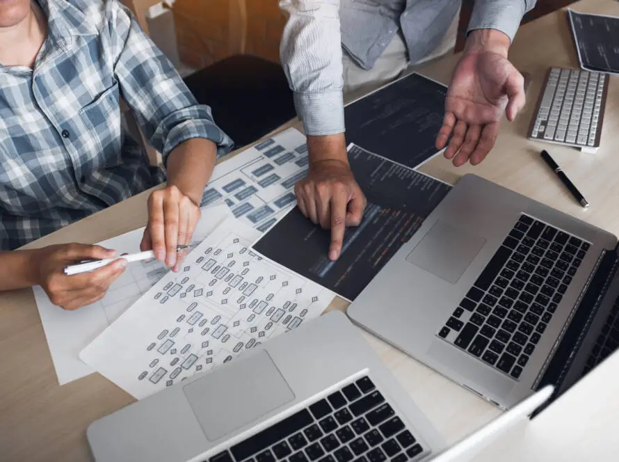 Two people work at a desk with laptops, printed charts, and code on paper. One points at code whilst the other holds a pen over a flowchart—suggesting digital marketing healthcare or software project planning. Hands and tools are visible, but not faces.