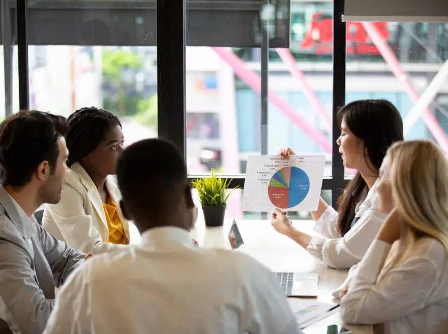 Four people sit around a table in a modern office as a woman presents a colourful pie chart—insights from their trusted medical marketing agency. A potted plant decorates the table, and large windows brighten the space with natural light.