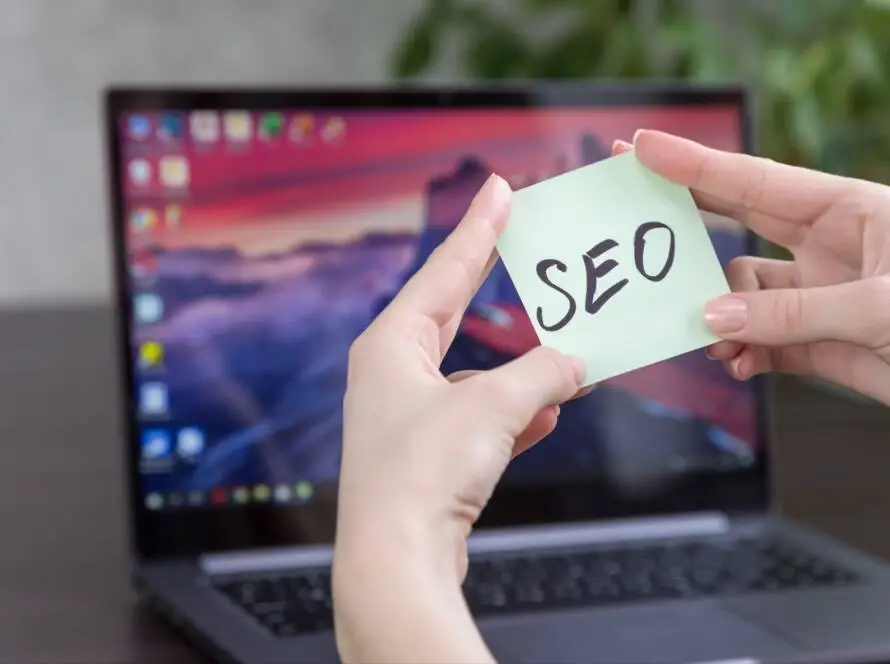 A person holds a light green sticky note with “SEO” written in bold black letters in front of an open laptop displaying a colourful, blurred desktop background; a green plant is visible—a nod to digital marketing healthcare.