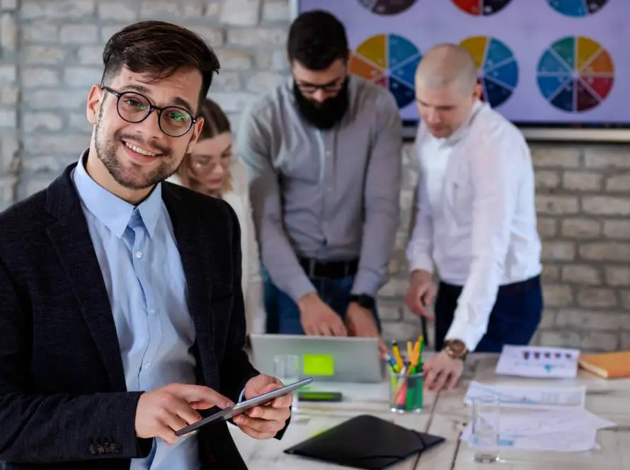 A smiling man in glasses and a suit holds a tablet in the foreground. Behind him, three colleagues from a healthcare digital marketing agency review documents and a laptop. A colourful pie chart decorates the brick wall in this modern office setting.