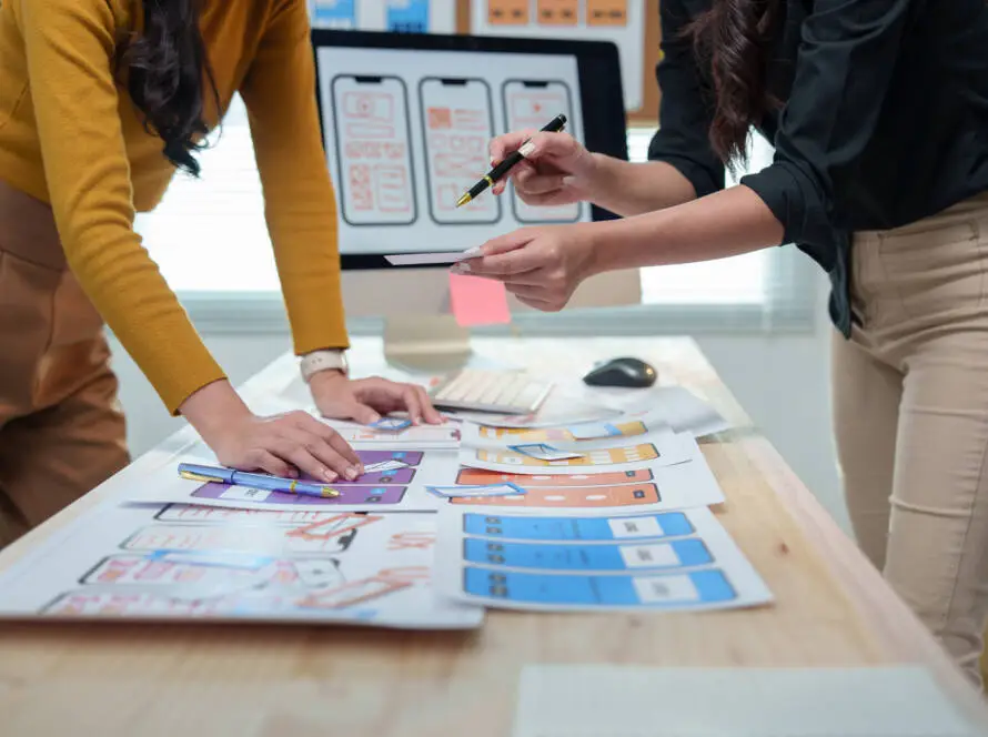 Two people stand at a desk covered with design mock-ups and colour samples, collaborating on UI wireframes for a healthcare digital marketing agency, suggesting a focused session on creative medical marketing solutions.