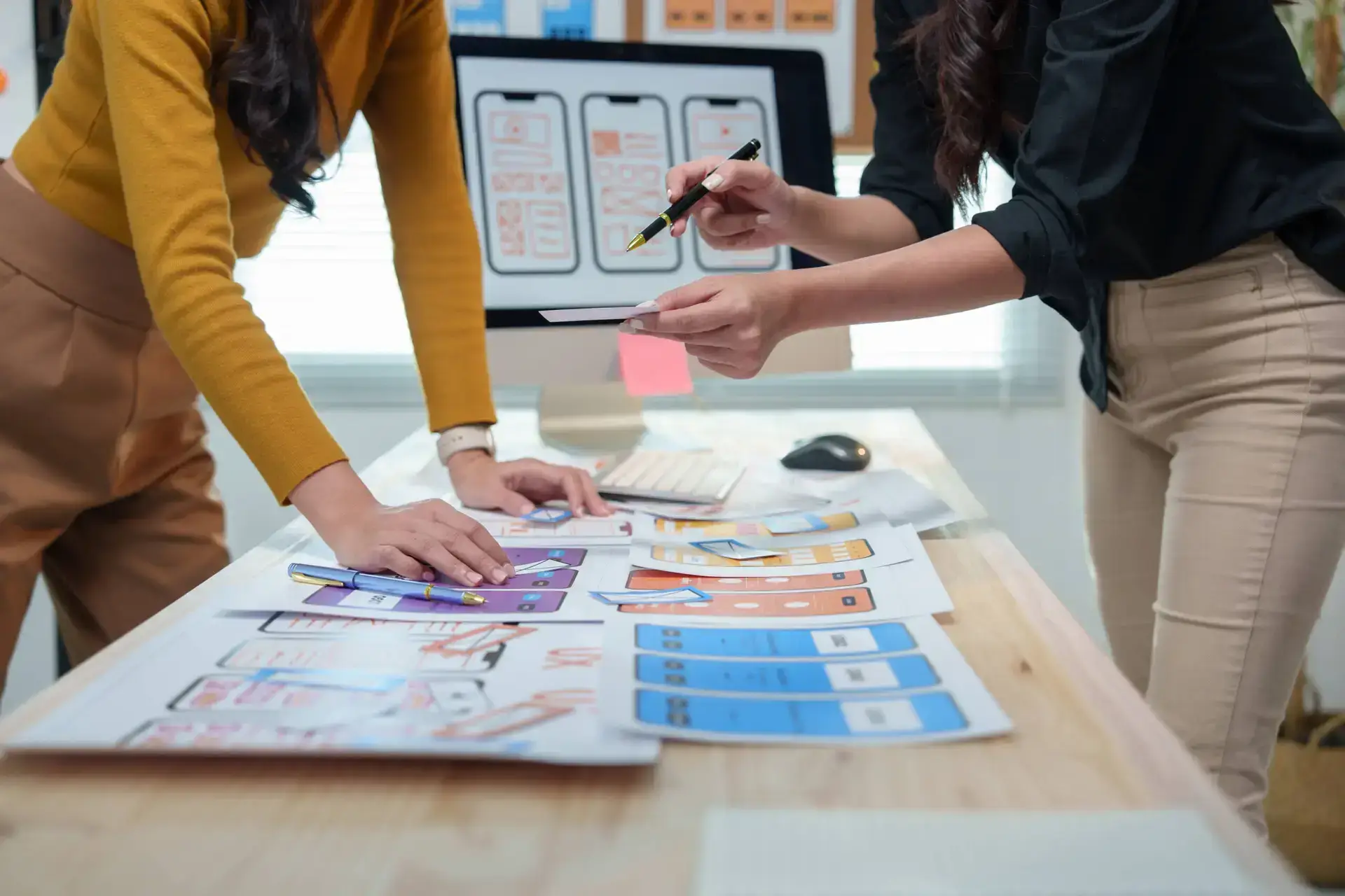 Two people stand at a desk covered with design mock-ups and colour samples, collaborating on UI wireframes for a healthcare digital marketing agency, suggesting a focused session on creative medical marketing solutions.