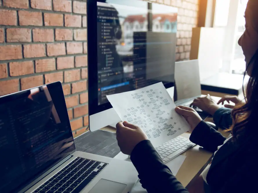 Two people sit at desks with laptops and large monitors displaying code. One person, from a medical marketing agency, holds a printed flow chart while examining the screen. The workspace features a brick wall and soft natural light from the right side.