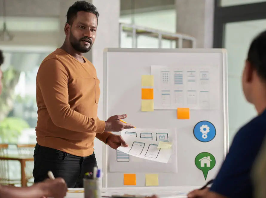 A man in a brown jumper stands by a whiteboard with diagrams and colourful sticky notes, leading a presentation on healthcare SEO to two colleagues. He gestures to papers with sketches, discussing innovative strategies in a modern office setting.