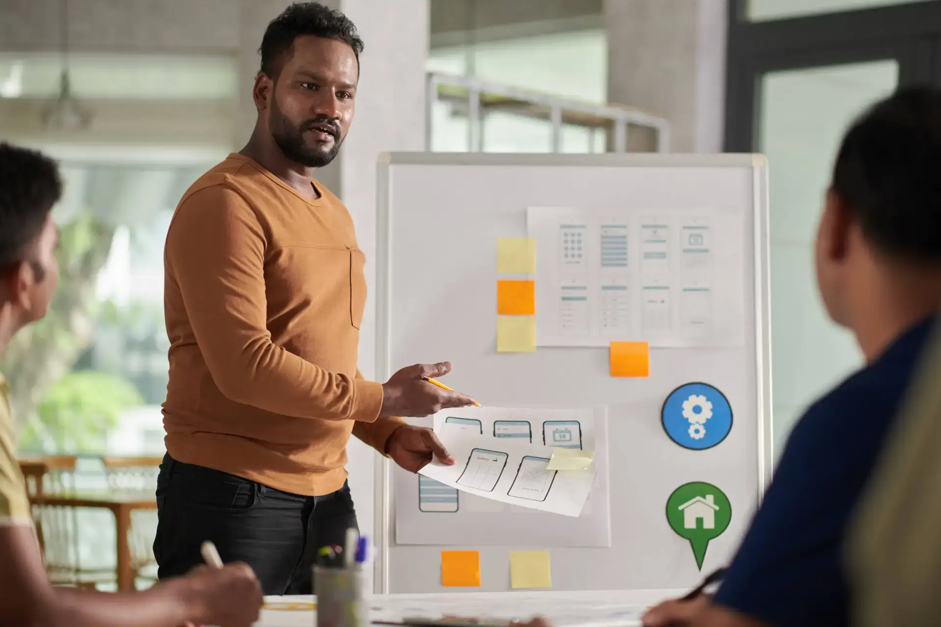 A man in a brown jumper stands by a whiteboard with diagrams and colourful sticky notes, leading a presentation on healthcare SEO to two colleagues. He gestures to papers with sketches, discussing innovative strategies in a modern office setting.