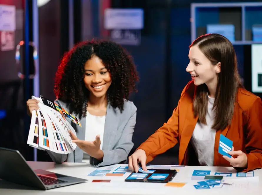 Two women sit at a desk in a modern office, discussing colour swatches and design samples. With a laptop open and materials spread out, they collaborate on branding ideas for a healthcare digital marketing agency’s latest creative project.