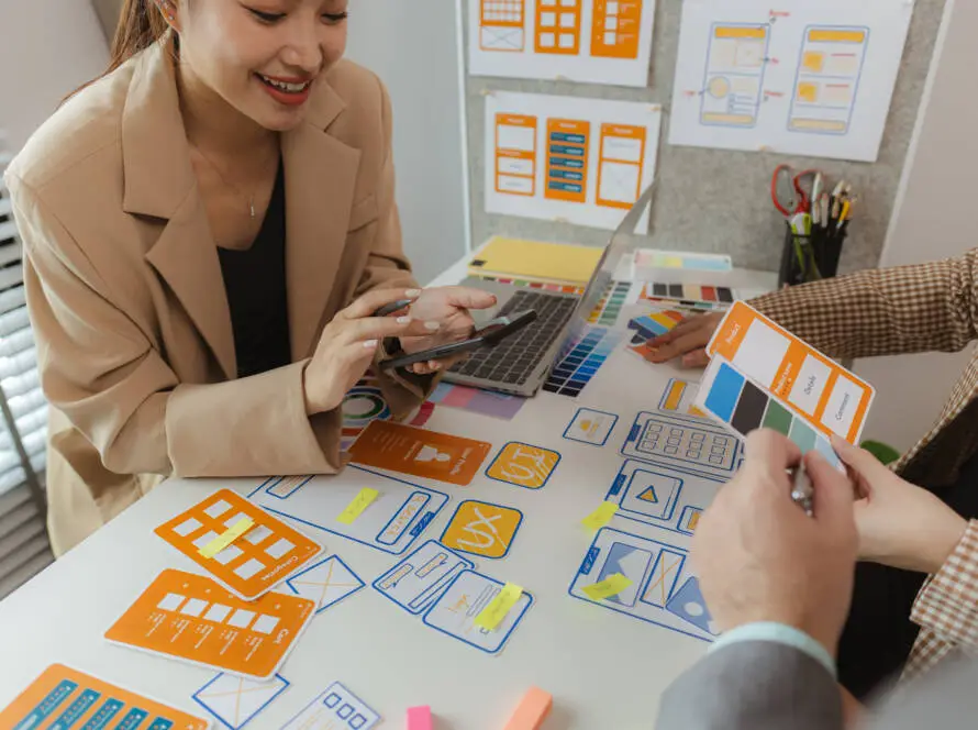 Two people sit at a desk covered with UX design mock-ups, colour swatches, and sticky notes. One smiles whilst holding a phone; the other selects a colour card—brainstorming innovative ideas for digital marketing healthcare campaigns.