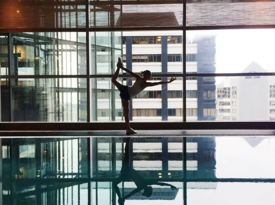 A person in a yoga pose stands on one leg by an indoor pool, holding one foot behind with an arm extended forward. Their reflection appears in the still water—a tranquil scene where a healthcare SEO consultant might find inspiration. Large windows reveal urban scenery.