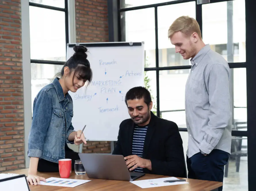 Three people stand around a table in a modern office, discussing work with a healthcare SEO consultant whilst looking at a laptop. A flipchart behind them displays a marketing plan. Papers, a red coffee mug, and a pen are on the table.