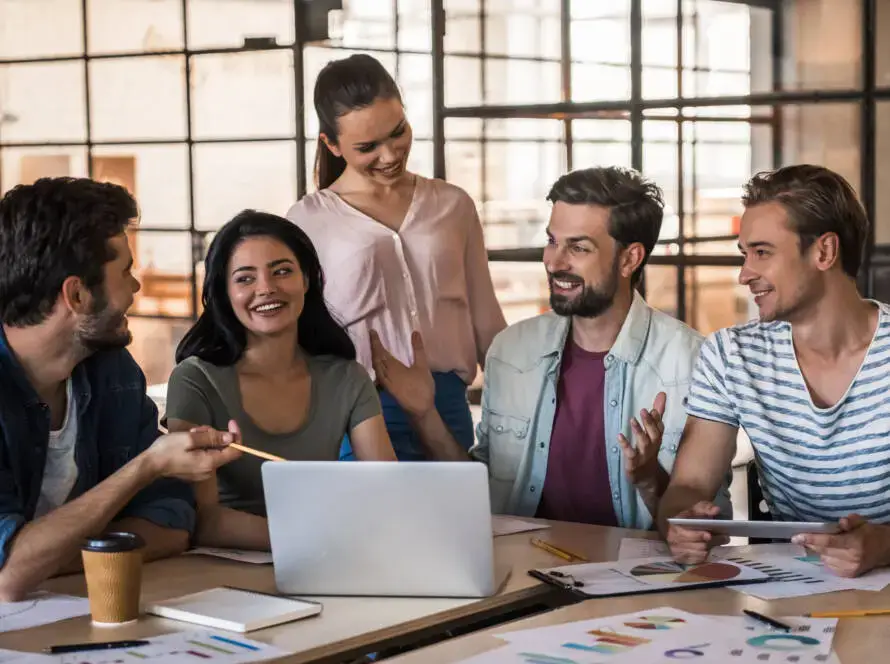 Five young adults sit and stand around a table with a laptop, papers, and coffee, engaged in lively conversation. They appear to be collaborating at a healthcare digital marketing agency in a modern office filled with light.