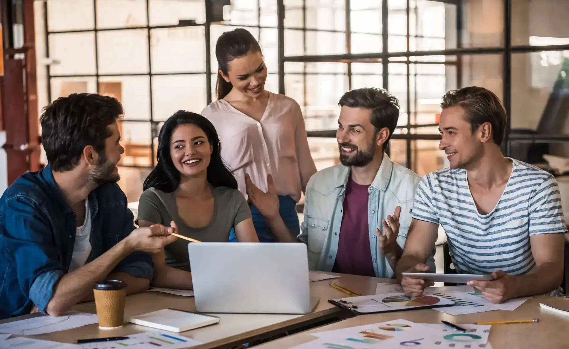 Five young adults sit and stand around a table with a laptop, papers, and coffee, engaged in lively conversation. They appear to be collaborating at a healthcare digital marketing agency in a modern office filled with light.