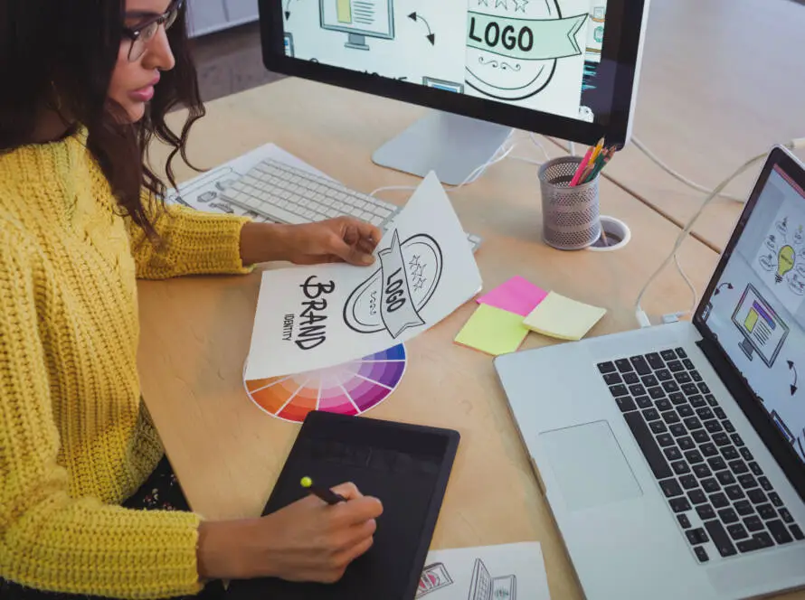 A woman in a yellow jumper sits at a desk with a laptop, graphics tablet, and colour samples. She holds a sheet labelled Brand with logo sketches for a healthcare marketing agency. Computer monitors show logo designs; sticky notes and pencils are nearby.