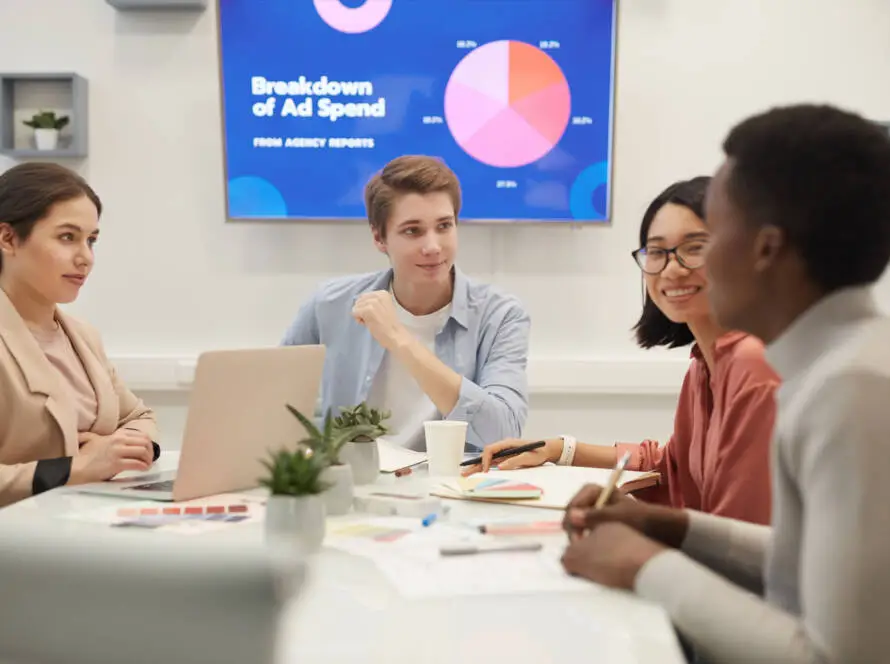 Four people sit around a table in a meeting room, discussing and smiling. Laptops, papers, plants, and a screen showing pie charts titled “Breakdown of Ad Spend from Agency Reports” reflect their focus on healthcare digital marketing agency strategies.