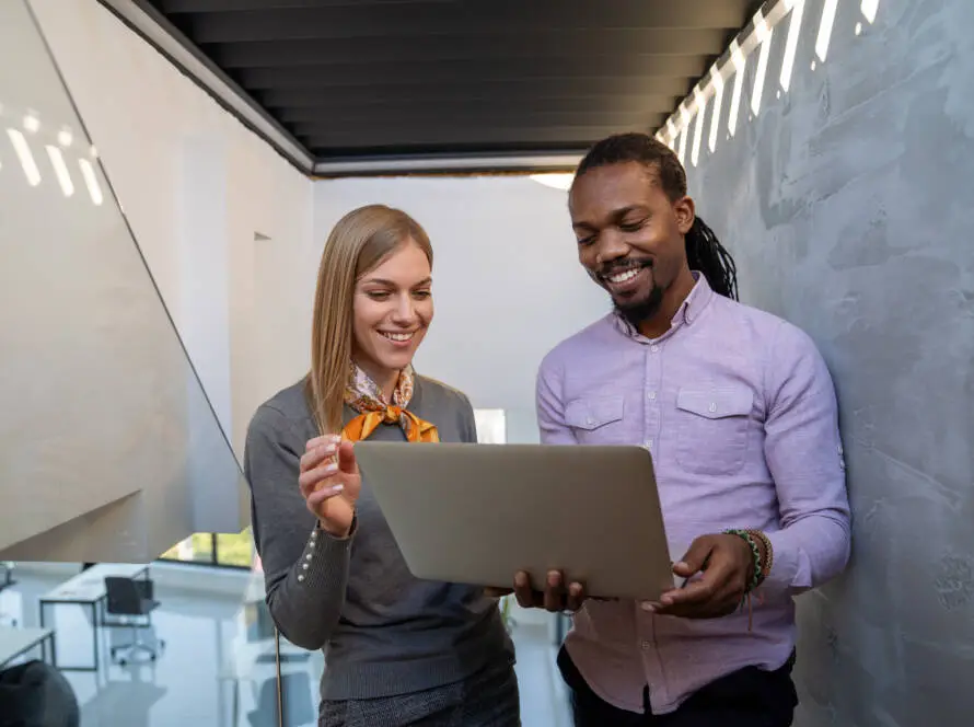 Two people stand indoors in a modern office, collaborating on a project for a healthcare digital marketing agency. The woman holds a pen, wearing a grey jumper and scarf, while the man in a light purple shirt smiles at the laptop screen.