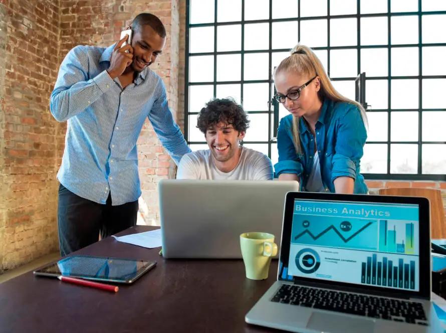 Three colleagues work in a modern office. A man talks on the phone while another smiles at a laptop as a woman stands beside him. The table, set for a healthcare digital marketing agency, holds business analytics, a yellow mug, notebooks, and a pen.