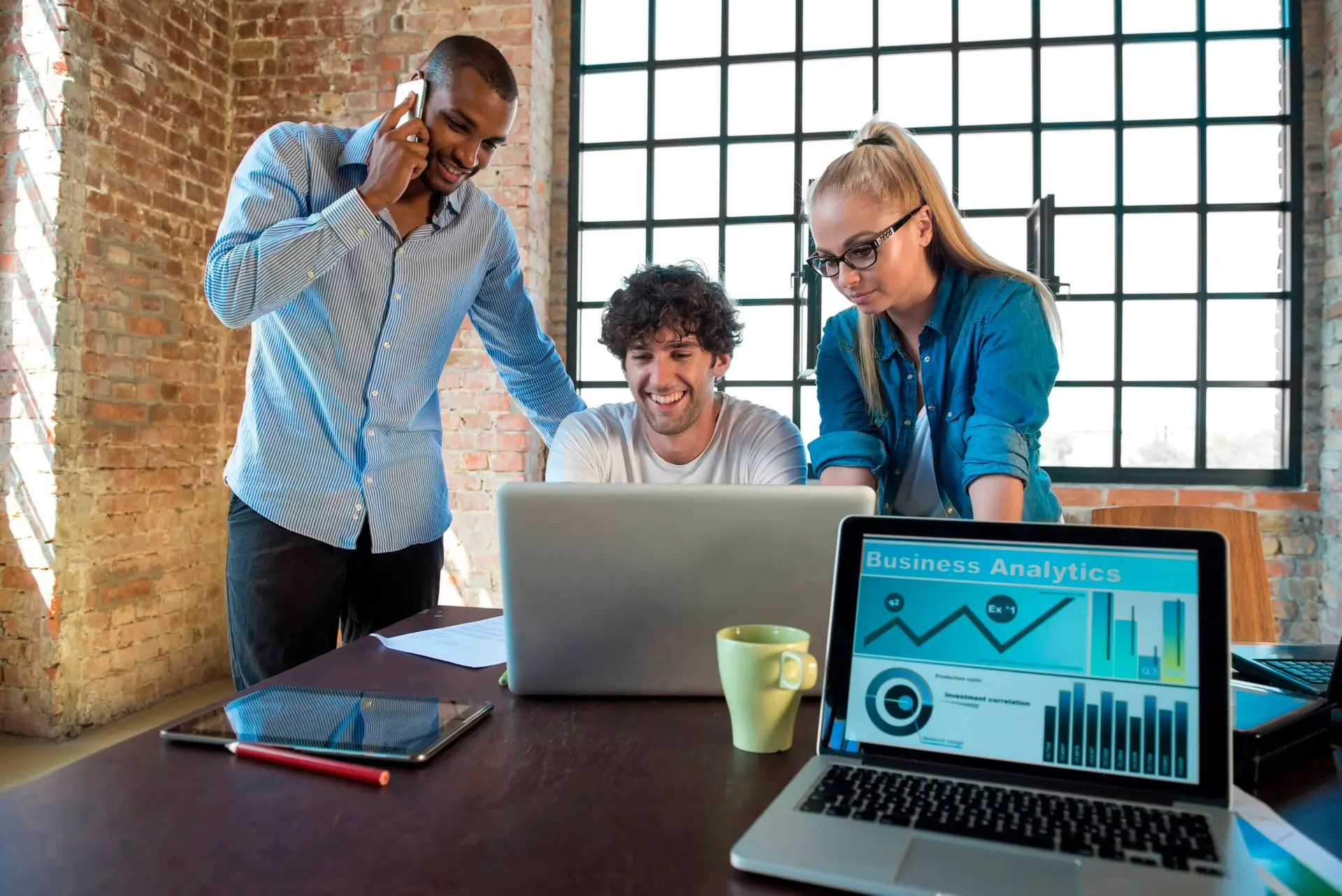 Three colleagues work in a modern office. A man talks on the phone while another smiles at a laptop as a woman stands beside him. The table, set for a healthcare digital marketing agency, holds business analytics, a yellow mug, notebooks, and a pen.
