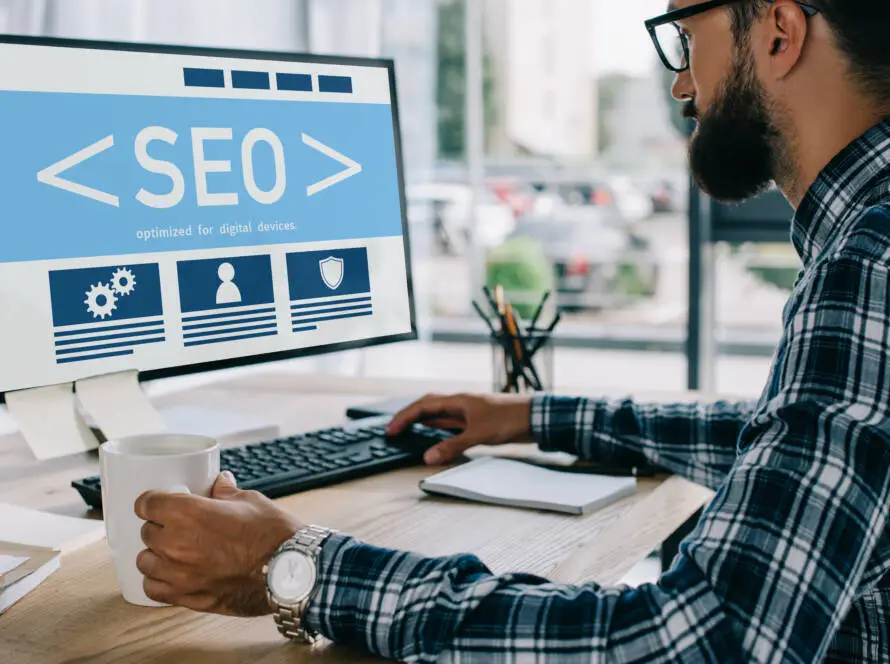 A man with a beard and glasses sits at a desk holding a coffee mug, working on a computer displaying “SEO” graphics—an ideal setting for a healthcare SEO expert or medical SEO agency specialising in digital marketing healthcare solutions.